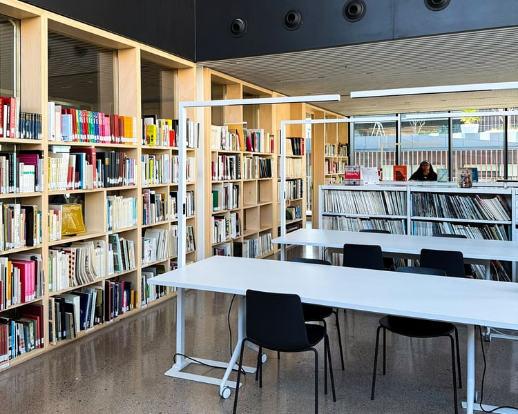 Die moderne Bibliothek der LCI Barcelona mit hohen Holzregalen voller Design- und Kunstbücher. Weiße Arbeitstische mit schwarzen Stühlen bieten Platz zum Lernen unter minimalistischen Langfeldleuchten, während im Hintergrund eine Person an einem weiteren Regal arbeitet.